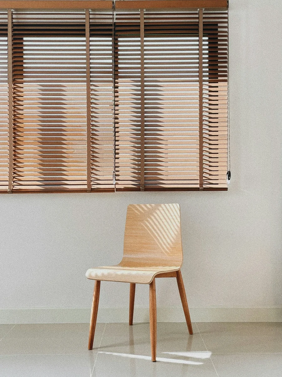 Wooden slat blinds half-lowered over a wide window above a single plywood chair