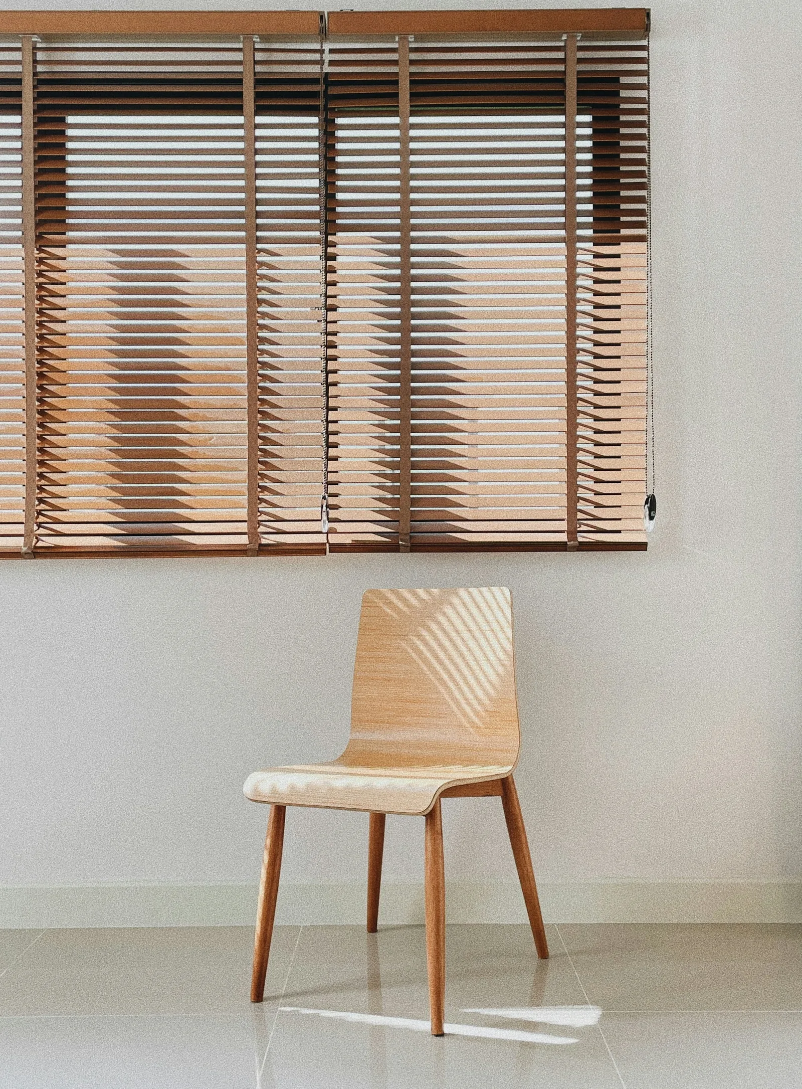 Wooden slat blinds half-lowered over a wide window above a single plywood chair