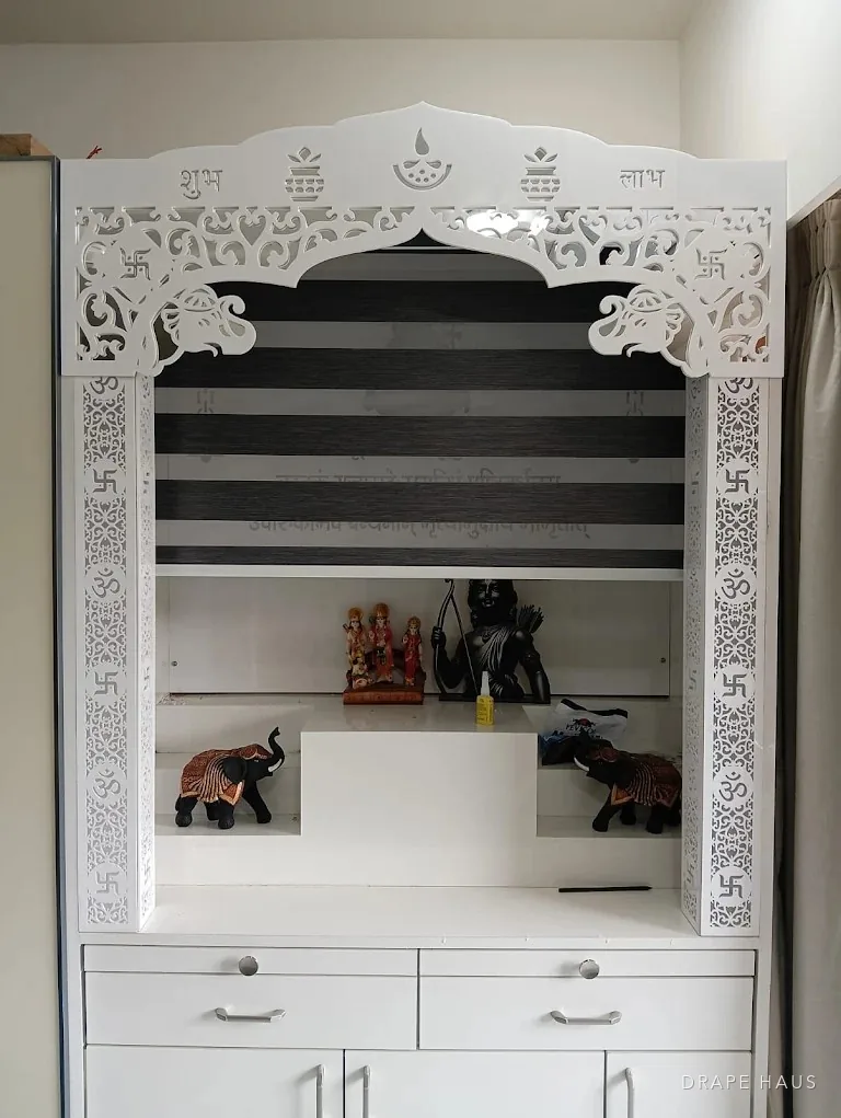 A white carved mandir unit with a charcoal zebra blind behind the arch, idols and two brass elephants on the shelves