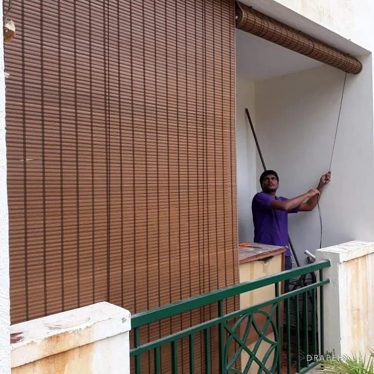 Bamboo chik blinds being installed across a first-floor veranda with a green metal railing