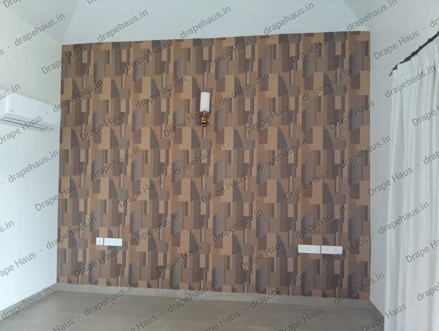 A bedroom wall papered in a brown and bronze geometric block pattern, with a brass wall light, two switch plates and a sheer curtain to the right