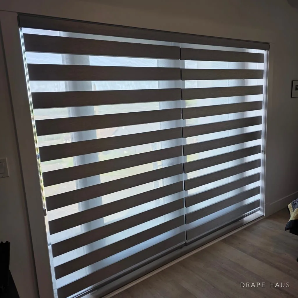 Two dark zebra blinds side by side covering a wide glazed opening onto a garden